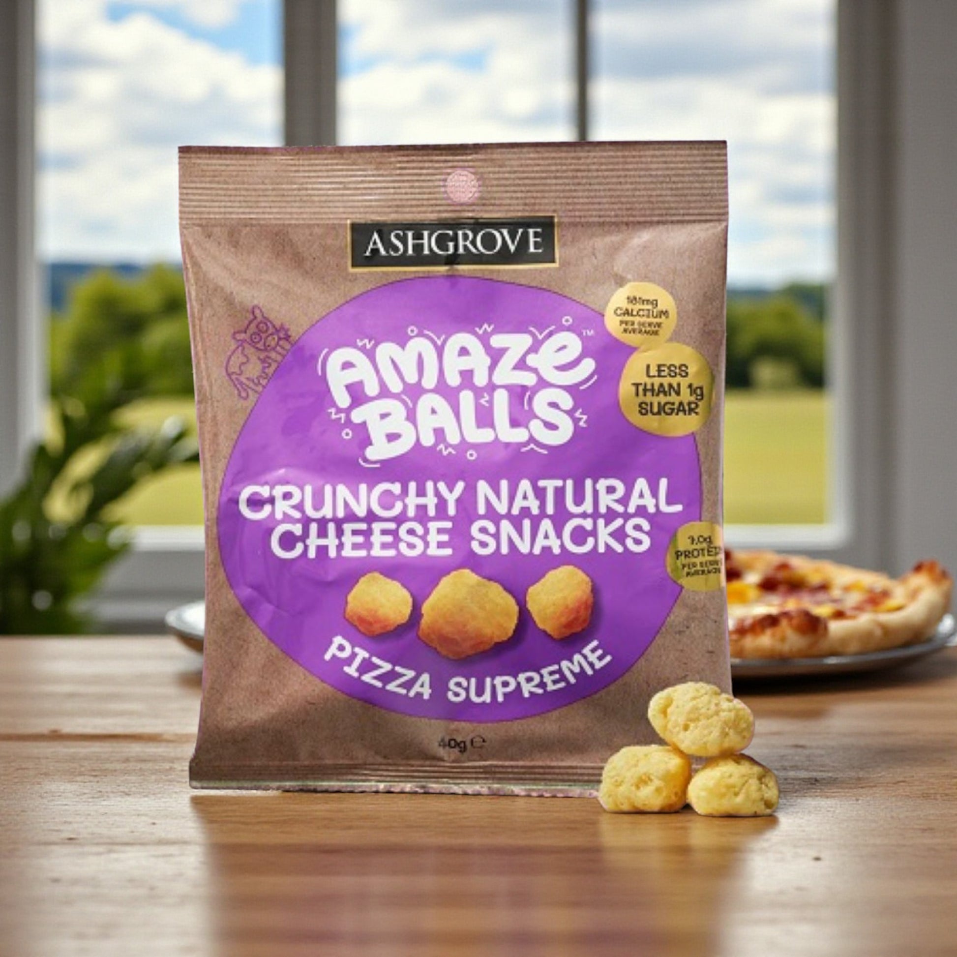 Packaging of Ashgrove Cheese AmazeBalls Crunchy Natural Cheese Snacks on a table with a pizza and a blurred outdoor background.