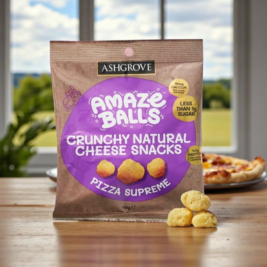 Packaging of Ashgrove Cheese AmazeBalls Crunchy Natural Cheese Snacks on a table with a pizza and a blurred outdoor background.