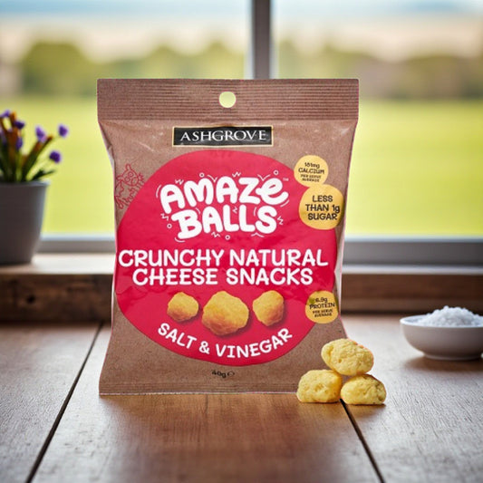 Packaging of Ashgrove Cheese AmazeBalls Crunchy Natural Cheese Snacks on a wooden surface with a blurred outdoor background.