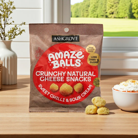 Packaging of Ashgrove Cheese Amaze Balls Crunchy Natural Cheese Snacks on a wooden table with a bowl of sour cream and sweet chilli in the background.