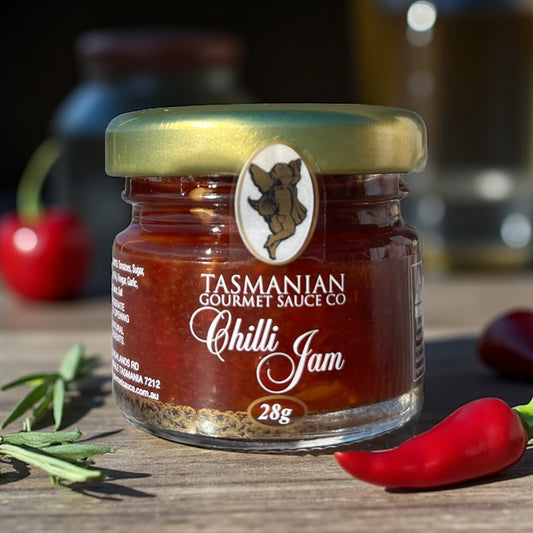 A glass jar of Tasmania Chilli Jam with a gold lid, placed on a wooden board with a green leaf and red chilli in the background.