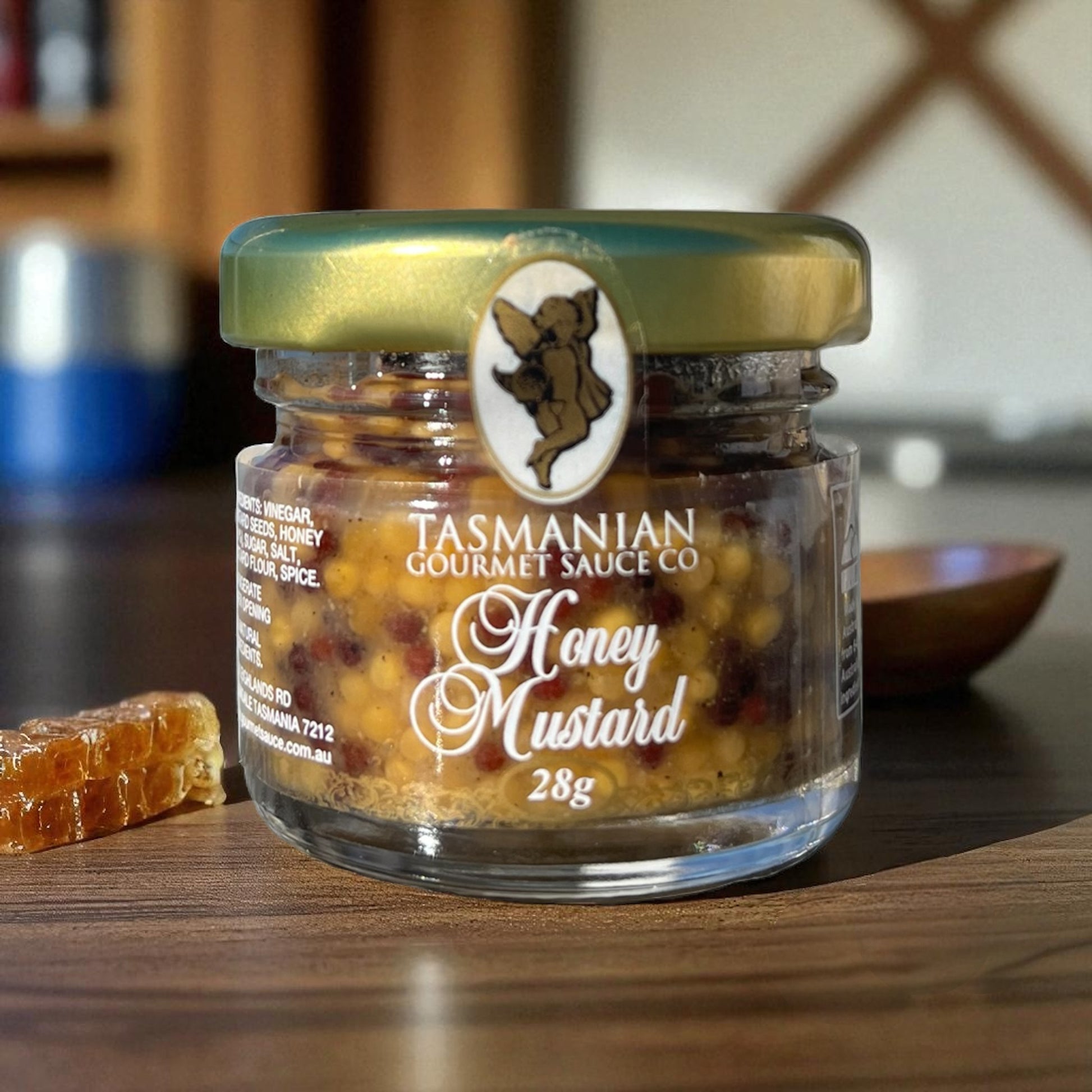 A jar of Tasmanian Honey Mustard sauce with a weight of 28g, placed on a wooden surface with a piece of honeycomb.