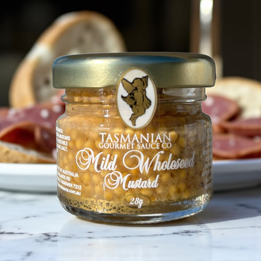 A glass jar of Mild Wholeseed Mustard by Tasmanian Gourmet Sauce Company, with a label showing the brand and product name, placed on a marble surface with bread and meats.