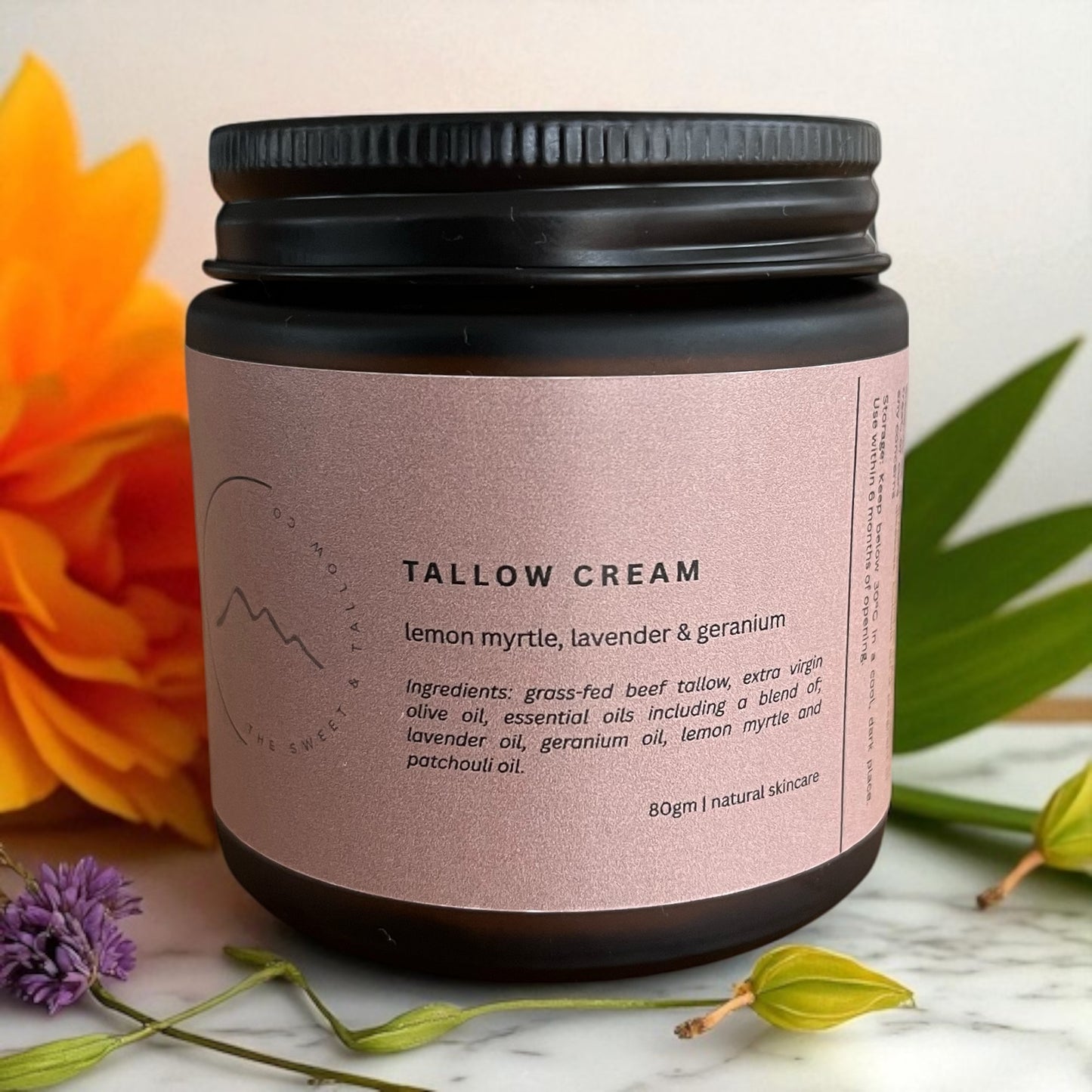 Jar of Tasmanian tallow cream with a pink label on a marble surface with flowers and leaves.