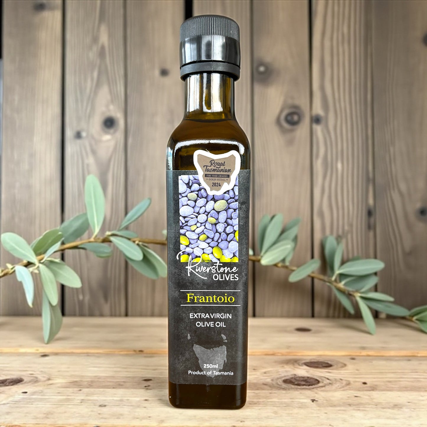 Bottle of Riverstone Olives Tasmanian Frantoio Extra Virgin Olive Oil on a wooden surface with an olive branch.