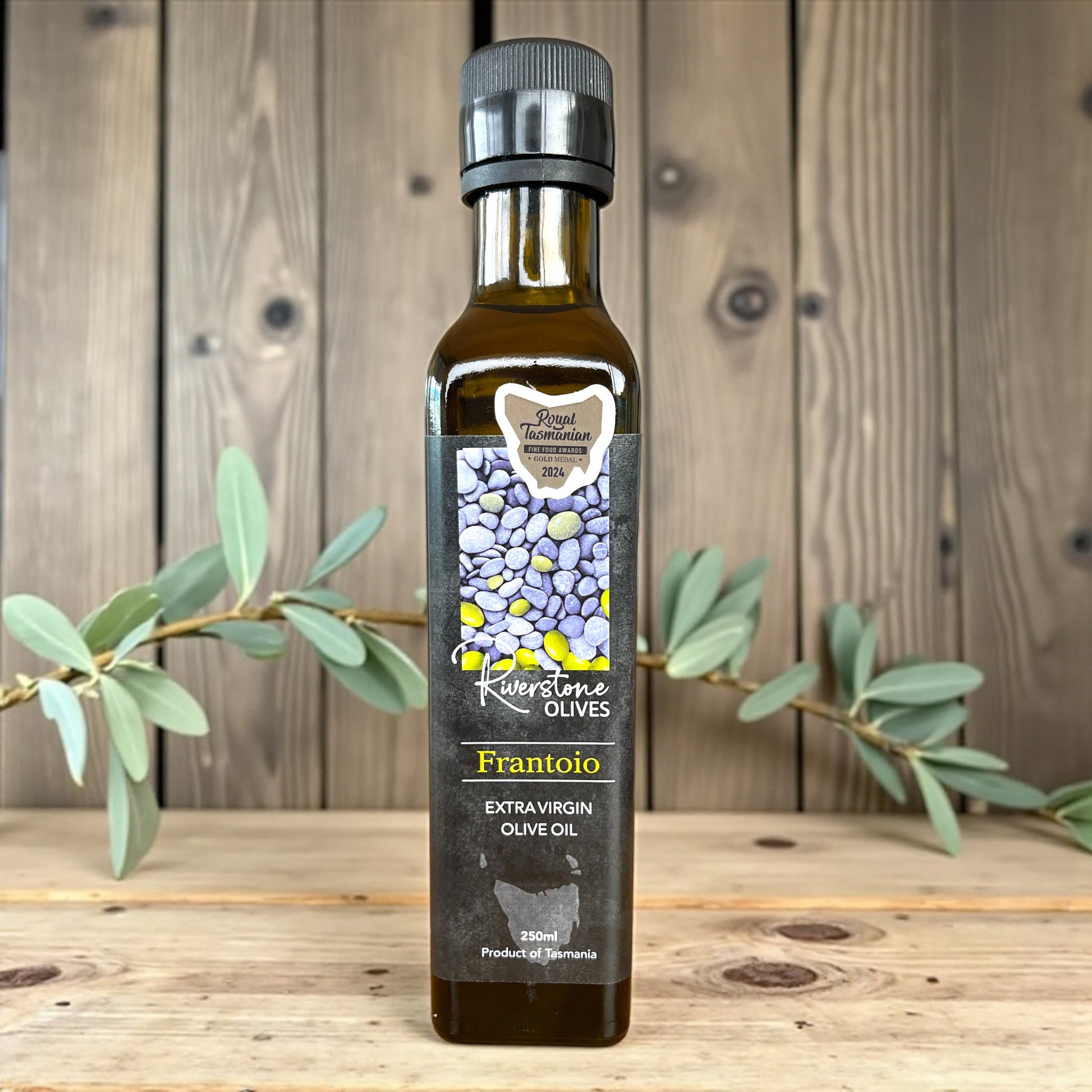 Bottle of Riverstone Olives Tasmanian Frantoio Extra Virgin Olive Oil on a wooden surface with an olive branch.