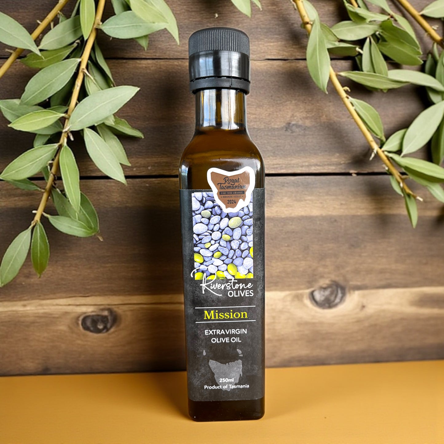 Bottle of Riverstone Olives Mission Tasmanian Extra Virgin Olive Oil on a wooden surface with olive branches.