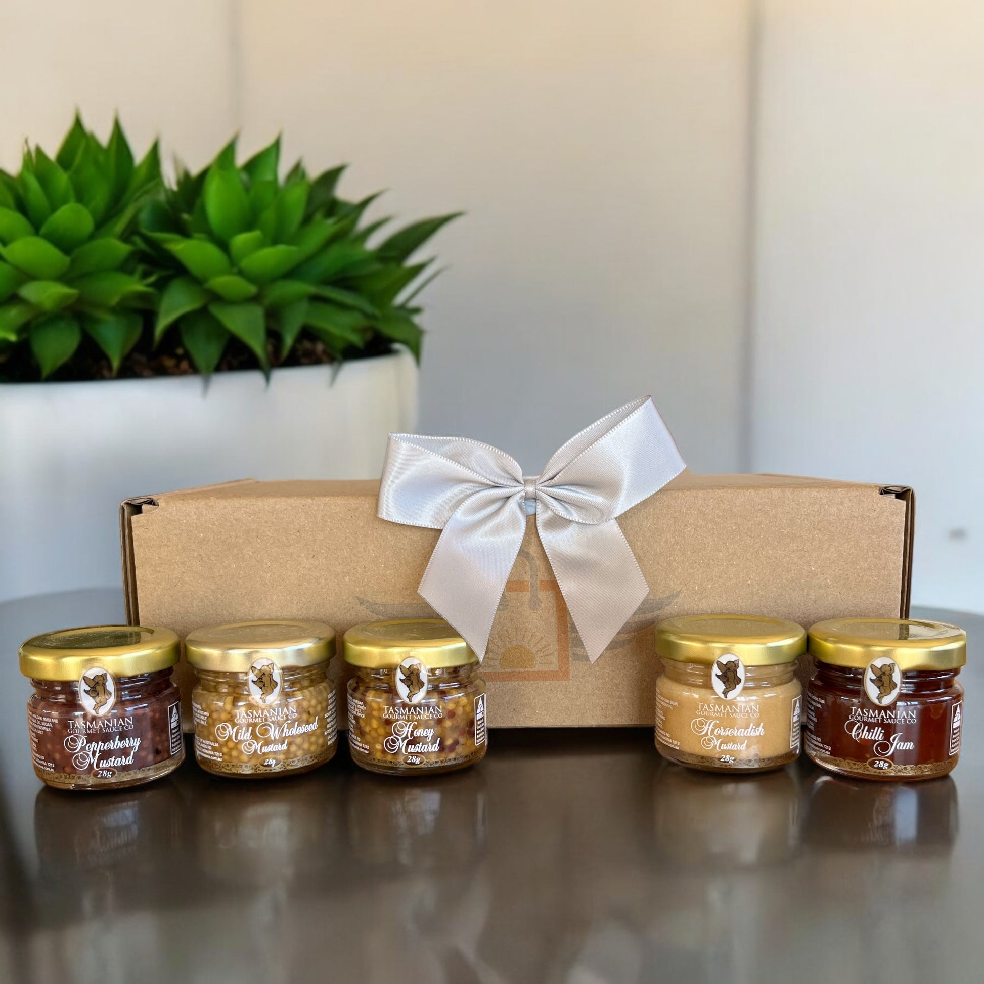 Five jars of Tasmanian gourmet mustards with a decorative box on a reflective surface, featuring a plant in the background.