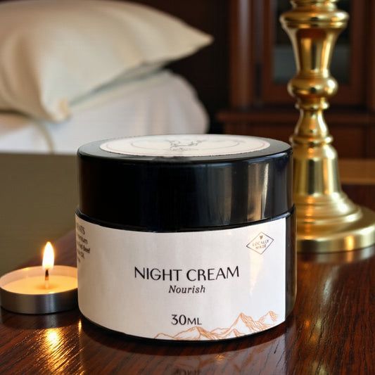 Jar of scented Tasmanian Tallow night cream on a wooden surface with a candle and lamp in the background