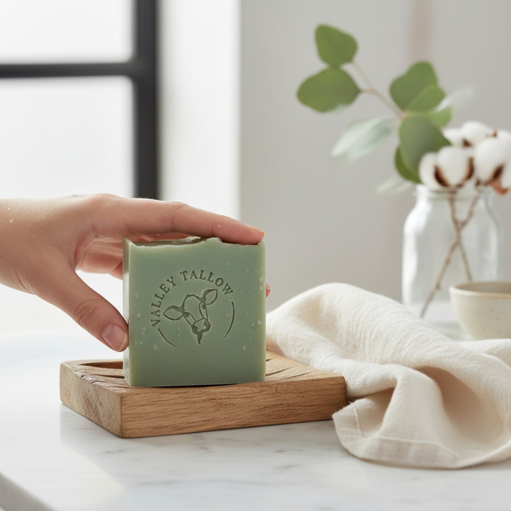 Hand holding Tasmanian Tallow Soap Lemongrass & Lime by Valley Tallow on wooden soap dish in a bright bathroom