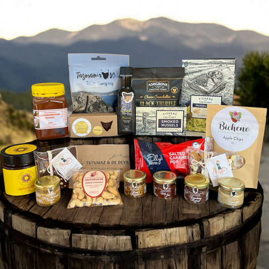 Tasmanian Gourmet Food Gift Hamper The Full Tassie gourmet food items including chocolate bars, olive oil, fruit jam, smoked mussels, nuts, honey, beef biltong, and more, displayed on a wooden barrel with a mountainous backdrop.